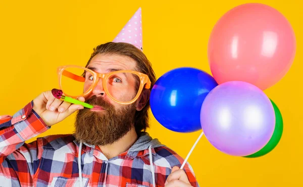 Bearded man in party hat and glasses with balloons blowing noisemaker. Birthday holiday celebration. — Stockfoto
