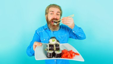 Sakallı adam yemek çubuklarıyla suşi yiyor. Japon yemek teslimatı. Tabakta zencefil ve suşi turşusu