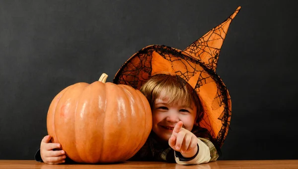 Child boy in witch hat with halloween pumpkin pointing to you. Trick or treat. Happy holidays