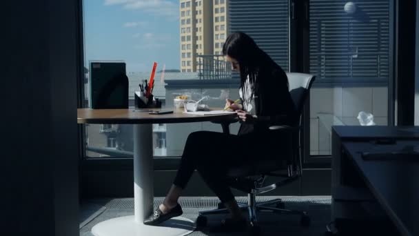 Zakenvrouw eet op werkplek.Jonge vrouw in een modern mooi kantoor in de buurt van het panoramische venster dineert en onderzoekt documenten — Stockvideo