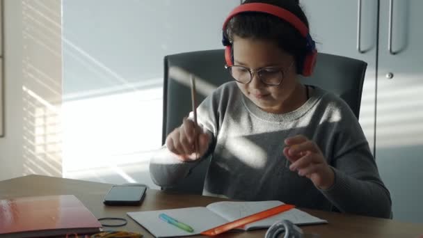 Criança bonita em fones de ouvido escreve em um caderno, faz lição de casa, estuda na mesa, distraindo-se jogando no telefone — Vídeo de Stock