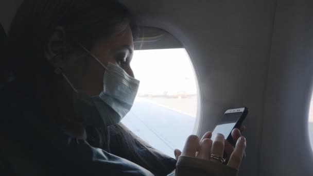Kobieta w masce używająca smartfona i wpisująca wiadomość przez telefon w samolocie przez okno podczas wieczornego lotu. Palce dotykają ekranu. Podróż w czasie trwania koronawiru — Wideo stockowe