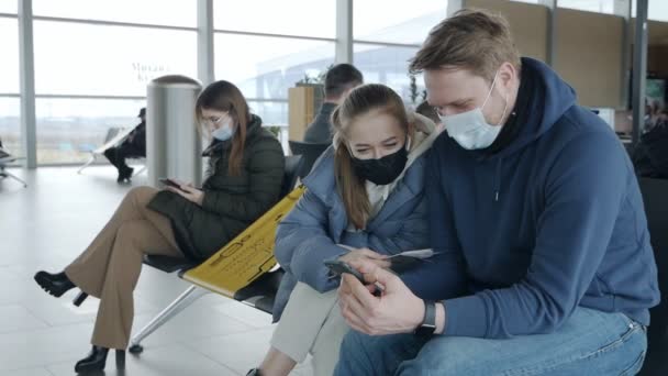 Mladí turisté v ochranných maskách sedí v letištní hale a dívají se na obrazovku telefonu. Cestovní koncept během koronaviru, společenská vzdálenost — Stock video