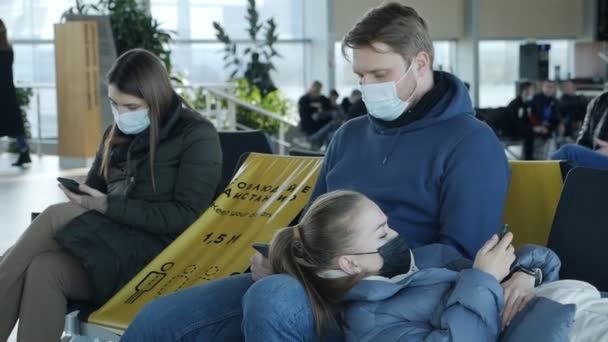 Una joven pareja con máscaras se sienta en un salón del aeropuerto. La chica está cansada y se acuesta en su regazo. Viaje durante la epidemia — Vídeos de Stock