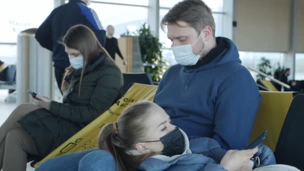 Молода пара в масках сидить в залі для аеропортів. Дівчина втомилася і лежить на колінах. Подорожі під час епідемії — стокове відео