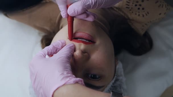 Närbild av en permanent make-up mästare med en röd penna ritar en kontur skiss av framtida läppar. Läppmikropigmentering, rodnad och tatuering, estetisk medicin tillvägagångssätt koncept — Stockvideo