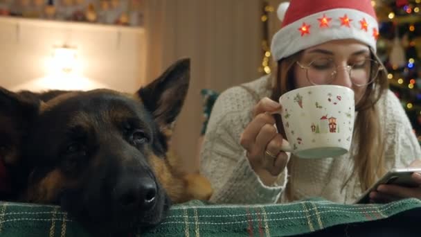 A woman lies on the couch near the New Year tree and looks at the phone screen, drinks coffee. Her dog is next to her. Cozy Christmas Eve in your apartment — Stock Video