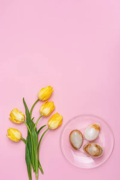 透明なプレートと春の黄色の新鮮な花の装飾イースターの卵コピースペースとピンクの背景にチューリップ ハッピーイースターカード フラットレイアウト トップビュー 垂直形式 — ストック写真