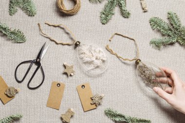 Kurumuş çiçekleri, otları dolduran incik boncuklar. Kırsal kumaş arka planında cam Noel süsleri. Kadınların elinde yeni yıl hediyesi ya da dekorasyon iç mimari fikri, xmas botanik baloları var.