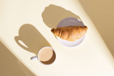 Fresh croissant and cup of hot coffee with milk in bright sunlight. Breakfast on summer morning