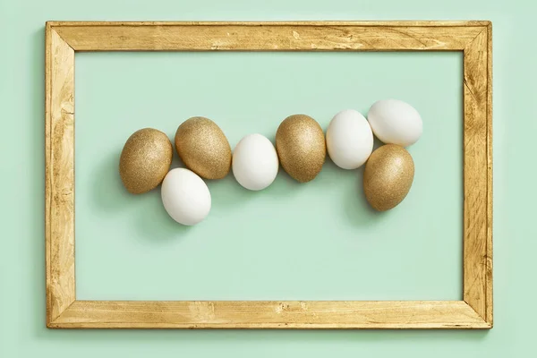 Pasen achtergrond met heldere eieren, glanzend goudkleurige en witte eieren op groen papier in houten frame. — Stockfoto