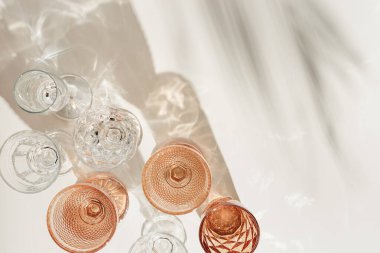 Şeffaf ve bej bardaklardan renkli bardaklar, güneş ışığında palmiye yaprağından güzel gölgeler, etkinlikler için yaz içeceği, beyaz şarap, moda bardaklarındaki su, estetik üst görünüm.