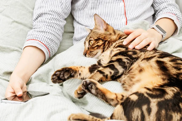 벵골 고양이와 함께 침대에 누워 잠옷을 입고 누워 있는 현대 소녀. 손님을 안고 있는 애완 동물. — 스톡 사진