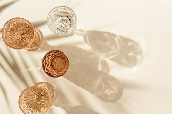 Набор цветных стаканов из прозрачного и бежевого стекла с красивыми тенями из пальмового листа при солнечном свете, летний напиток для мероприятий, белое вино, вода в трендовых стаканах, эстетический вид сверху Стоковое Фото