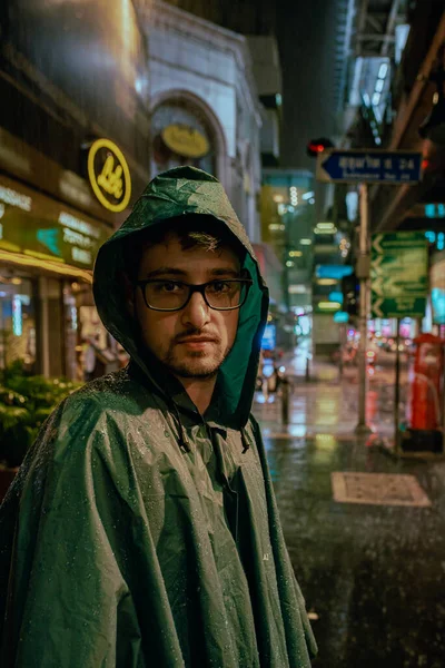 Man wearing a raincoat looks at camera in the monsoon rain on the streets of Bangkok.