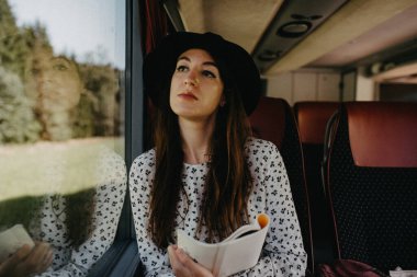 Elinde not defteri olan düşünceli genç bir kadın pencereden dışarı bakan bir otobüsle seyahat ediyor..