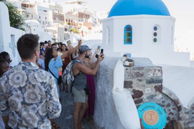 Oia, Yunanistan - 9 Eylül 2022: Oia 'da dar, kalabalık ve kalabalık caddeler, Santorini bir yaz öğleden sonra popüler Yunan Adası' nda bir kedinin fotoğrafını çeken akıllı telefonlu turistlerle dolu.