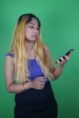Sarışın kadın yeşil arka planda dururken telefona bakıyor.