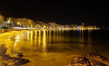 Geceleri plajda yanan binalar ve suda yansıyan ışıklar. Alicante Ara.