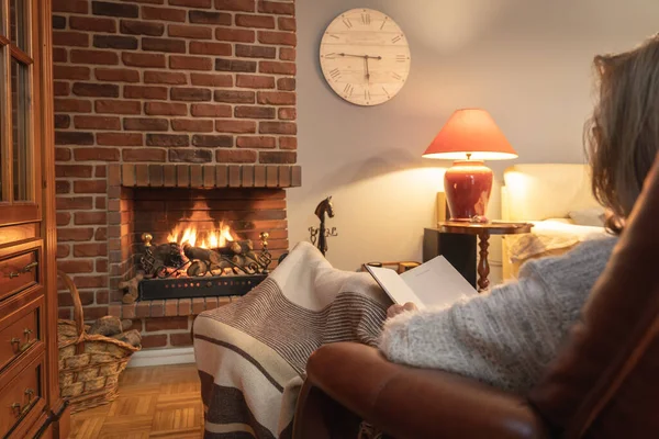 Woman reading a book in a relaxed way in front of the fire of a fireplace in winter.