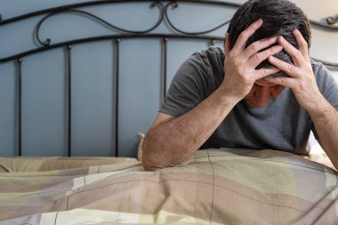 Uykusuzluk çeken bir adam ellerini başının üstüne koyup yatakta oturuyor ve yorgunluktan ölüyor..