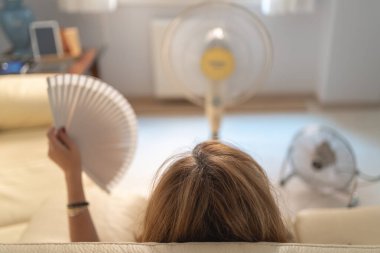 Woman sitting on a sofa with several fans giving her air and a fan in her hand, high summer temperatures