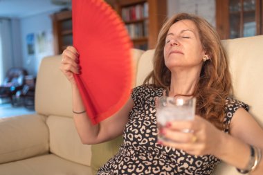 Woman giving herself air with a fan and a glass of water with ice to cool off in summer