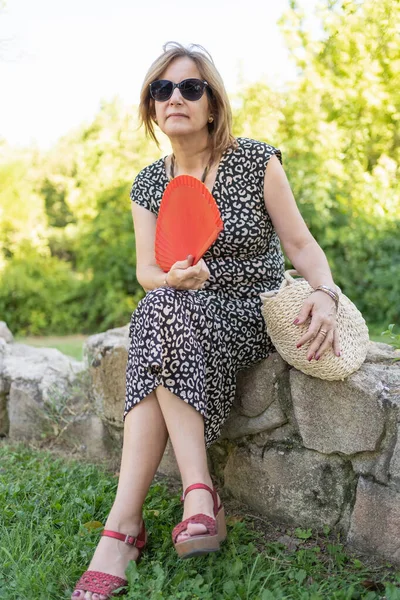 Mature woman with a fan and sitting in an outdoor park on a hot summer day