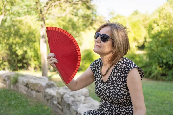 Woman with a lot of heat and giving herself fresh air with a fan in a public park