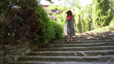 Woman walking down the stairs of a public park in a dress and straw hat on a sunny day
