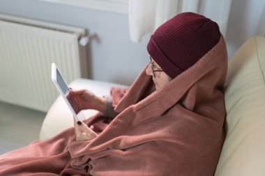 Battaniyeye sarılmış, yün şapkalı bir kadın soğuk kış ve enerji tasarruflarıyla ısınırken tablete bakıyor.