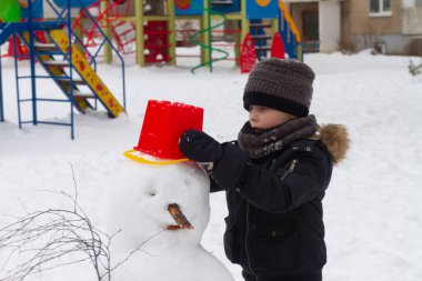 Kışın parkta bir çocuk kafasında kırmızı bir kovayla kardan adam yapar..