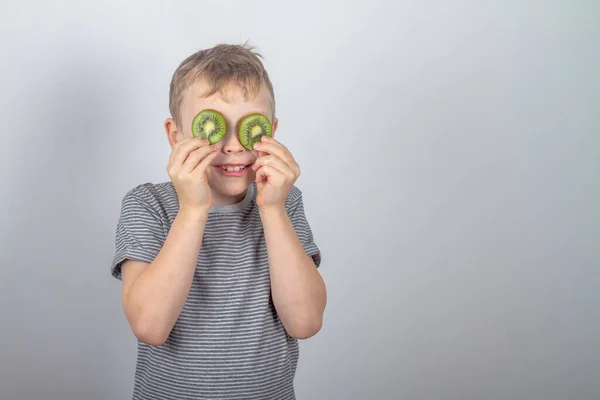 Wesoły biały chłopiec zamknął oczy plastrami zielonych owoców kiwi na szarym tle w studio — Zdjęcie stockowe