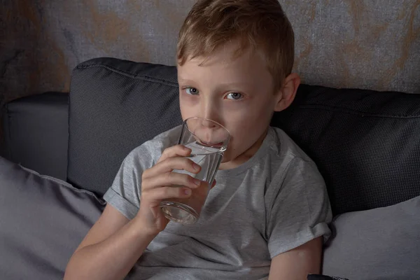 一个高加索男孩坐在灰色的沙发上喝着杯子里的水 — 图库照片