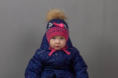 2 yaşında, beyaz bir kız çocuğu. Sıcak kış kıyafetleri içinde. Mavi tulum, şapka, pembe çizmeler ve gri arka planda eldivenler..