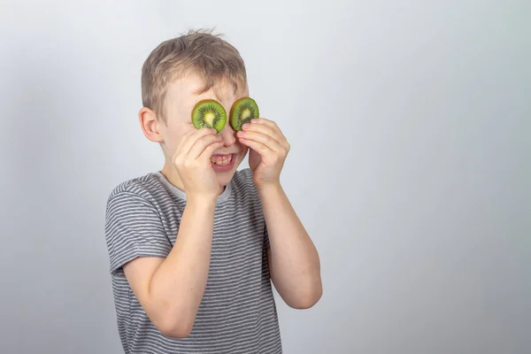 一个快乐的白人男孩在工作室的灰色背景上用几片绿色的猕猴桃水果闭上了眼睛 — 图库照片