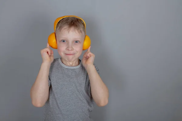 グレーのTシャツを着た金髪の7歳の白人少年とオレンジで作られたオレンジ色のヘッドフォンが架空の音楽を聴いて遊んでいます スペースのコピー — ストック写真