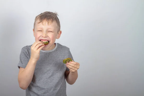 一个高加索男孩吃着一个绿色的猕猴桃 在灰色的背景上笑着 — 图库照片