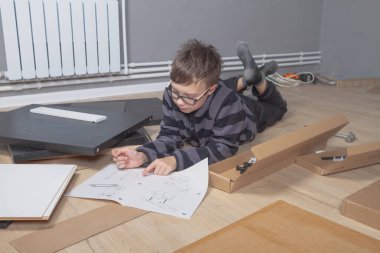 Genç bir çocuk yere uzanmış mobilya montajının talimatlarını okuyor..