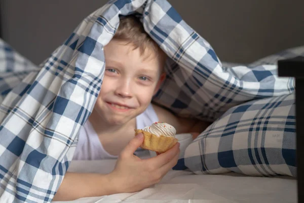 一个白人男孩正在床上躺在一个温暖的毛毯下吃着一个美味的奶油蛋糕 — 图库照片