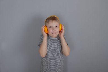 Gri tişörtlü ve portakaldan yapılmış turuncu kulaklıklı 7 yaşında sarışın bir beyaz çocuk hayali müzik dinliyor, çocuğun yüzünde neşeli bir duygusal ifade var. Boşluğu kopyala.