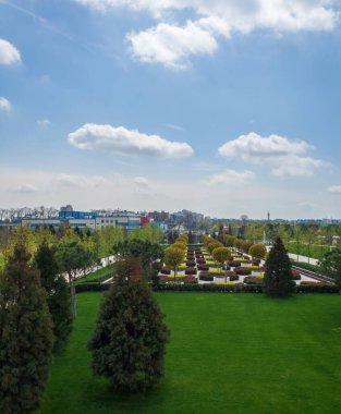 Mavi gökyüzünün altında beyaz bulutlu zarif bir şehir parkı. Baharın başında güzel bir parkın manzarası. Şehir manzarası. Sokakta ağaçlarla çevrili yeşil çimenler.