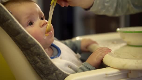 Κοντινό πλάνο πλάνα του μωρού που τροφοδοτείται με κουτάλι με πουρέ λαχανικών και κάθεται σε μια καρέκλα — Αρχείο Βίντεο