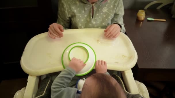 Baby sits in highchair in the kitchen and turn over the plate moving it on table filmed from above — Stock Video