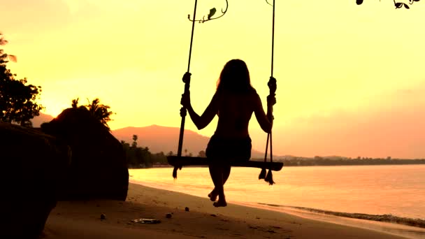 Задний вид молодой женщины, сидящей на качелях на пляже рядом с океаном на закате — стоковое видео