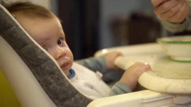 Küçük çocuk sandalyeye oturur ve annesinin ona verdiği ezilmiş meyveden püre yer.