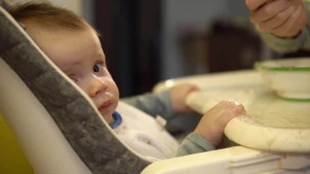 Il bambino siede su un seggiolone e mangia purea di purè di frutta che sua madre gli sta dando. — Video Stock