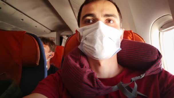 Joven hombre caucásico se sienta dentro del avión con máscara médica facial y almohada del cuello y mirando — Vídeos de Stock