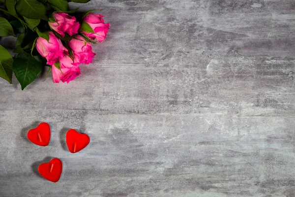 Szekrényfelvétel rózsaszín rózsákról és szív alakú vörös gyertyákról szürke fa felületen Stock Fotó