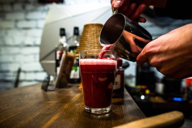Barmenin elleri ahşap tezgahta duran şeffaf camlara kırmızı kokteyl dökerken yakından görülüyor.